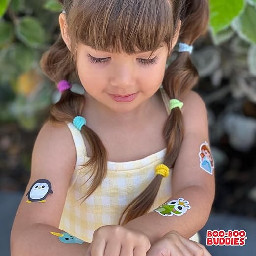 Smiling girl in yellow dress with Boo-Boo Buddies® bandages on arms featuring penguin, frog, narwhal, and princess characters – fun adhesive bandages for kids