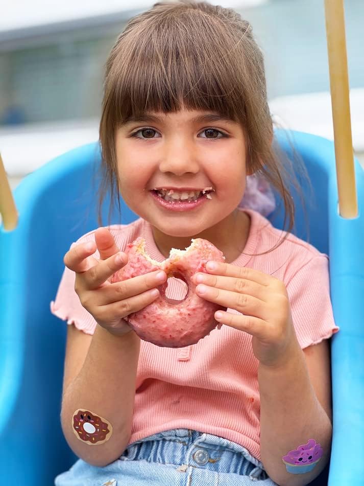 Girl smiling while eating pink donut with Boo-Boo Buddies® donut and cupcake bandages on her legs – fun dessert-themed bandages for kids