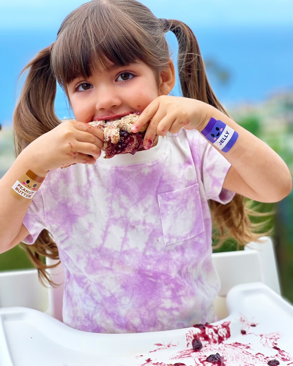 Girl eating messy sandwich wearing Boo-Boo Buddies® Peanut Butter and Jelly bandages on both wrists – cute food-themed bandages for kids with personality