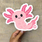 Hand holding a large Boo-Boo Buddies® collectible axolotl vinyl sticker with cute pink gills and smiley face against a neutral background
