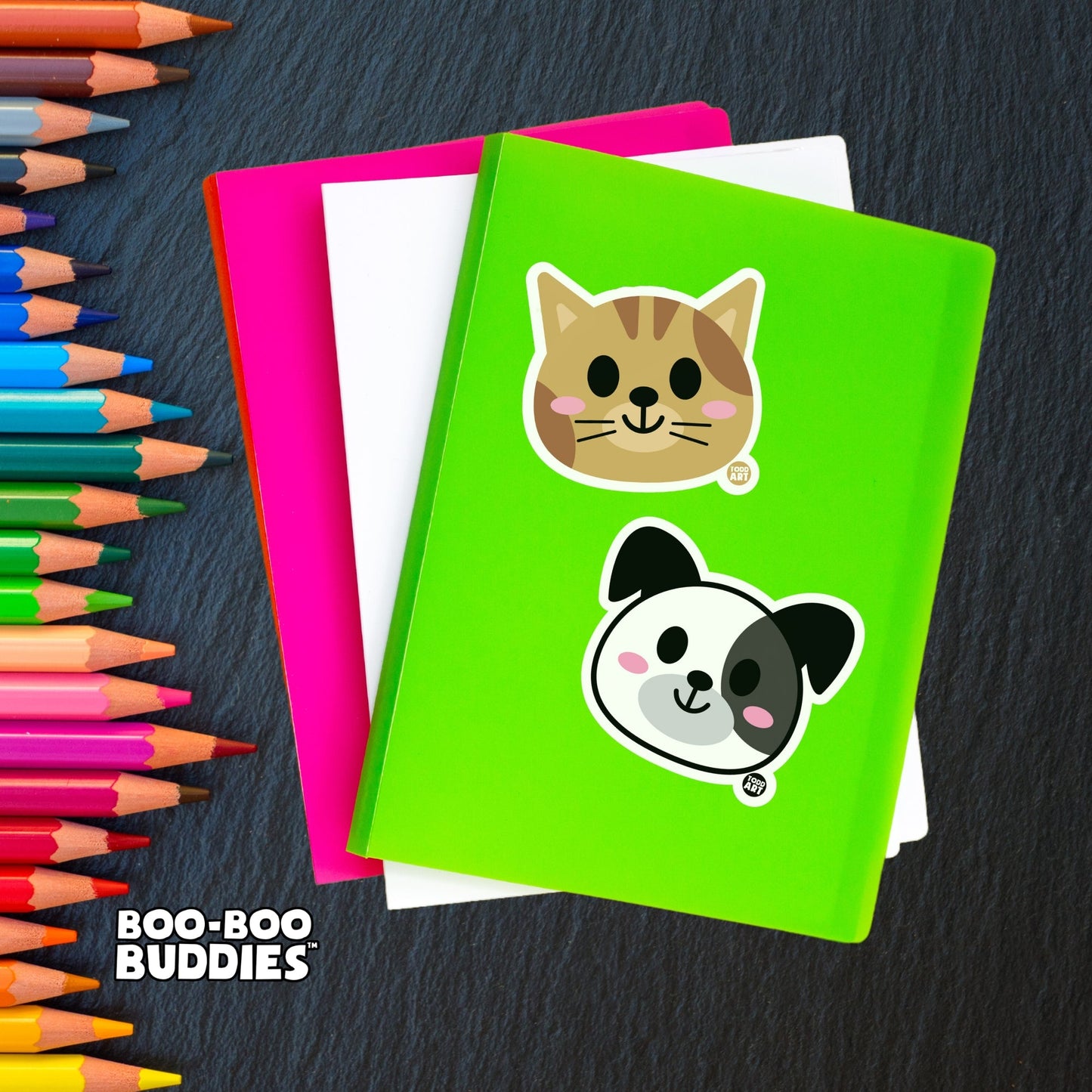 Notebook with a Boo Boo Buddies sticker surrounded by colorful pencils on a desk.
