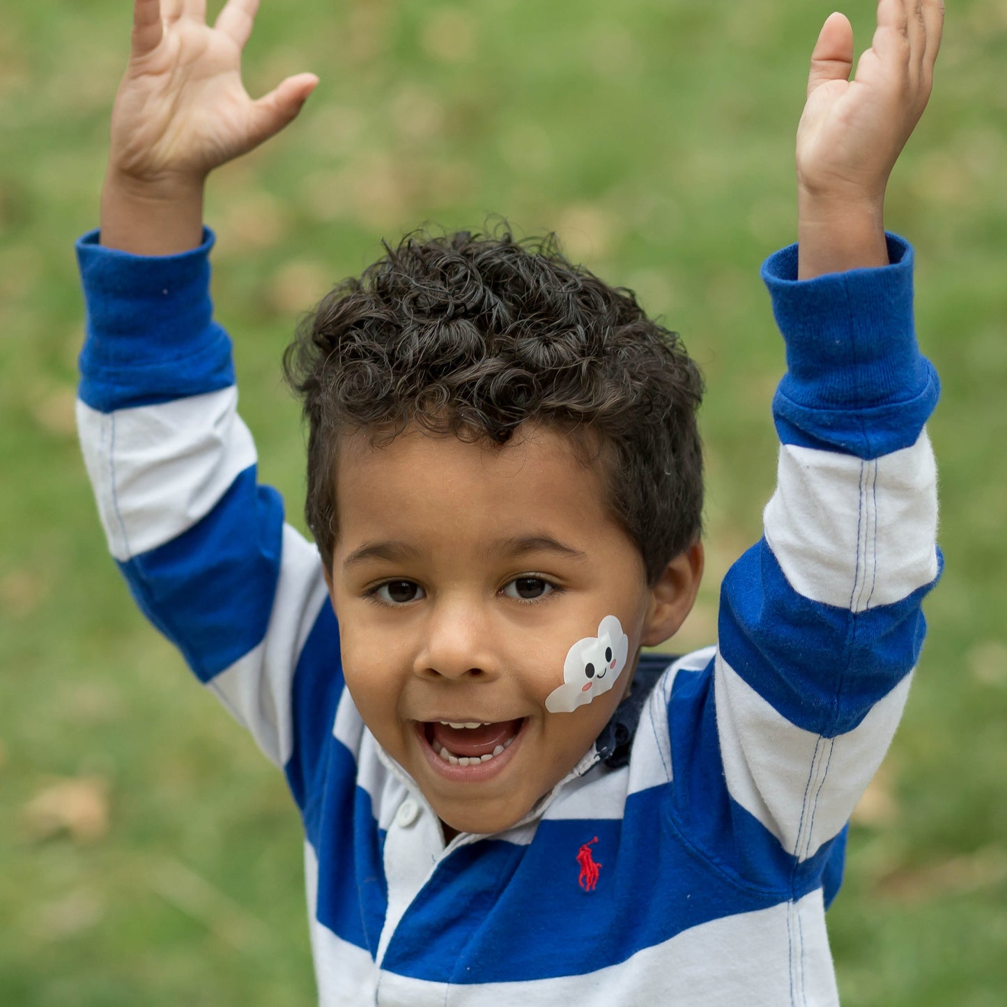 Smiling child raising his arms while wearing a Boo Boo Buddies fun and colorful kids bandage on his cheek outdoors.
